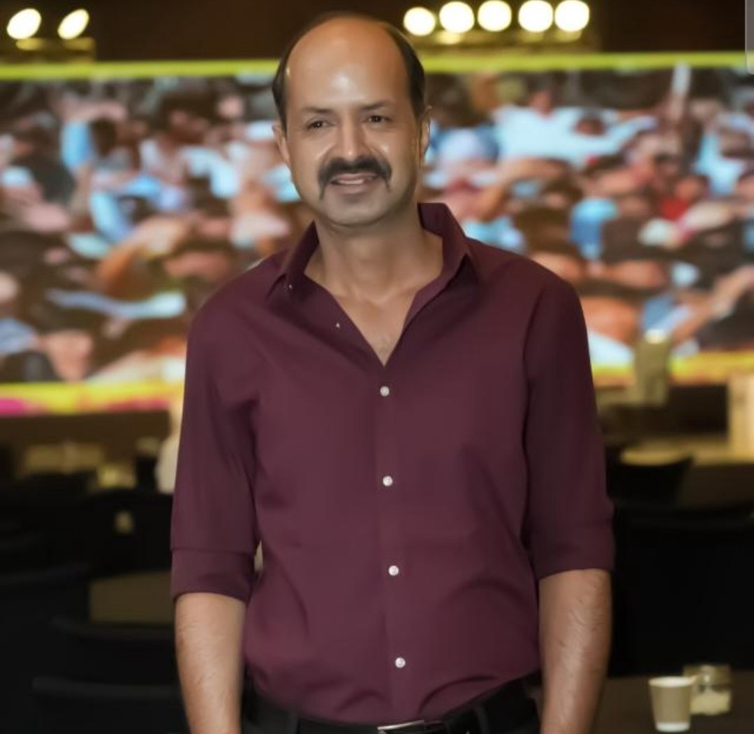 77d917b2-ac98-4646-b1df-21879fbc1bca | No. 1 Indian Media News Portal Man in a maroon button-down shirt posing for a photo with a blurred crowd in the background at an event.