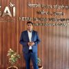 Man in a navy suit standing with hands folded in front, in a wooden-paneled lobby with 'SAI' and Netaji Subhas Western Centre text on the wall