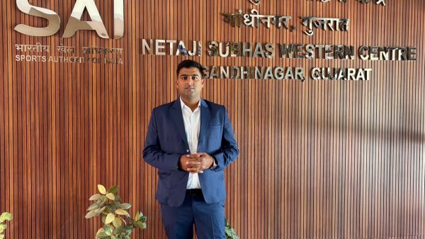 Man in a navy suit standing with hands folded in front, in a wooden-paneled lobby with 'SAI' and Netaji Subhas Western Centre text on the wall