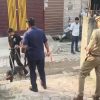 Uniformed police officer watches as two men hold a dog on leashes in a courtyard street near storefronts.