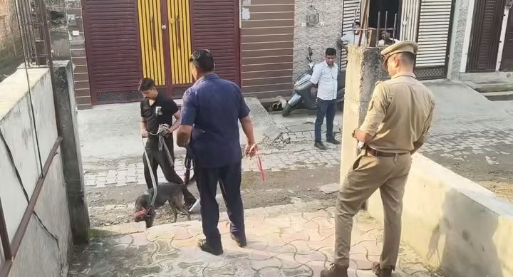 Uniformed police officer watches as two men hold a dog on leashes in a courtyard street near storefronts.