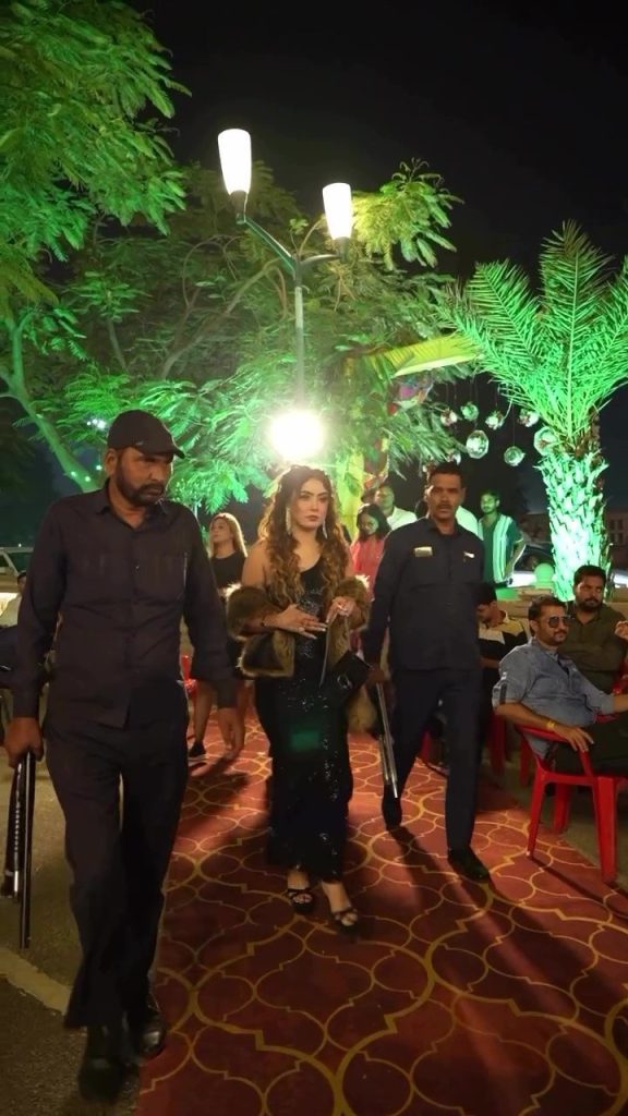 Woman in a black gown with a fur stole walks along a lit red-carpet path flanked by two security guards at a nighttime outdoor event.