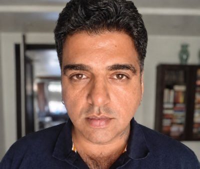 Close-up portrait of a man with dark hair wearing a navy polo shirt, indoors with a bookshelf in the background.