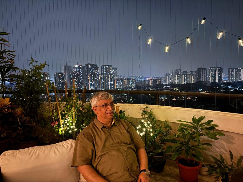 Sanjaya | No. 1 Indian Media News Portal Older man sits on a patio sofa at night on a rooftop garden, city skyline lit up in the background with string lights above.