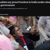 Female journalist with glasses being restrained by security while a microphone is pushed toward her in a crowded scene of reporters and police nearby.