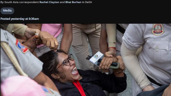 Female journalist with glasses being restrained by security while a microphone is pushed toward her in a crowded scene of reporters and police nearby.