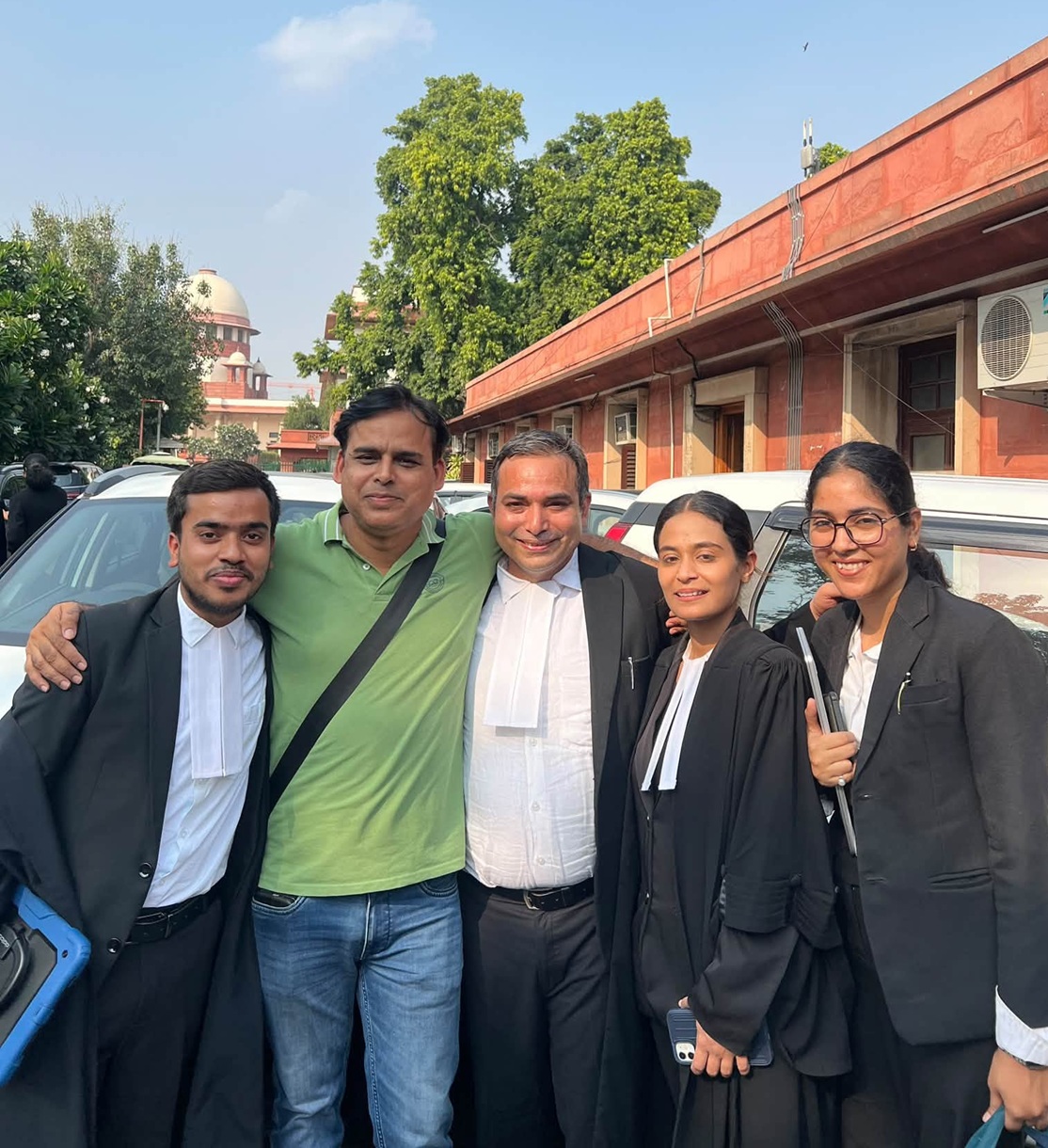 Five people pose together outdoors near parked cars; four wear black graduation gowns, one in a green polo shirt, smiling at the camera.
