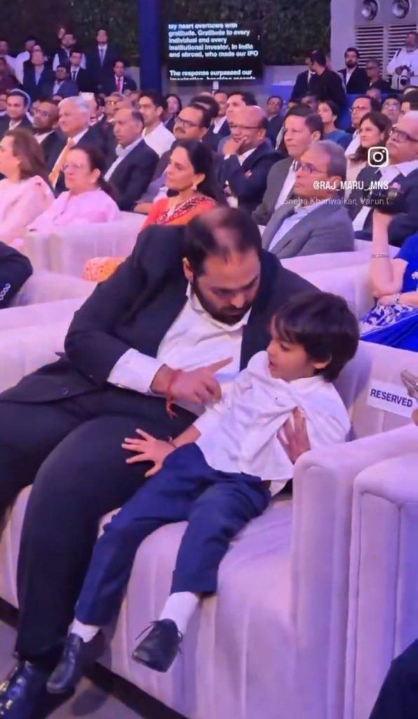 ambani_anant | No. 1 Indian Media News Portal A man in a dark suit sits in a crowded event row with a young boy on his lap, the boy touching the man's chest.