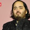 Portrait of a man with shoulder-length dark hair and beard, wearing a black traditional jacket, at a pink-background event with a Network18 logo.