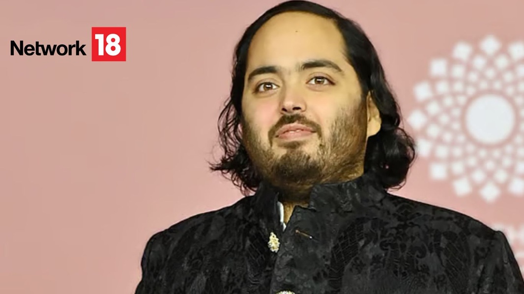 Portrait of a man with shoulder-length dark hair and beard, wearing a black traditional jacket, at a pink-background event with a Network18 logo.