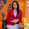 Smiling woman in a red blazer sits in a black chair at a glass desk in a world-map themed TV studio backdrop.