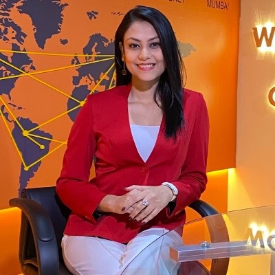 Smiling woman in a red blazer sits in a black chair at a glass desk in a world-map themed TV studio backdrop.