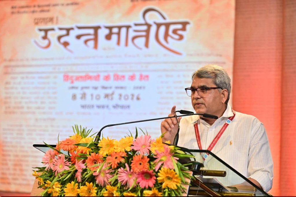 Man in glasses and a white shirt speaks at a podium decorated with pink, yellow, and orange flowers; a large banner is in the background.