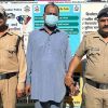 Three men stand outdoors: two police officers in khaki uniforms flank a man wearing a blue kurta and a face mask, in front of a public-safety information board.