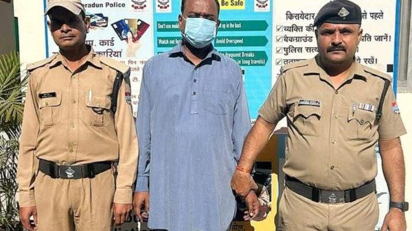 Three men stand outdoors: two police officers in khaki uniforms flank a man wearing a blue kurta and a face mask, in front of a public-safety information board.
