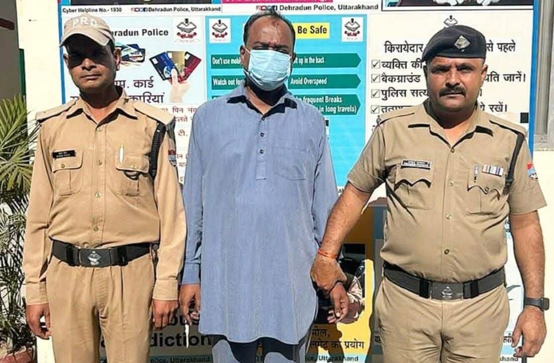 Three men stand outdoors: two police officers in khaki uniforms flank a man wearing a blue kurta and a face mask, in front of a public-safety information board.