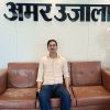 Man seated on a brown leather sofa in an office lobby; large Hindi text on the wall behind him.