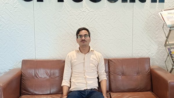 Man seated on a brown leather sofa in an office lobby; large Hindi text on the wall behind him.
