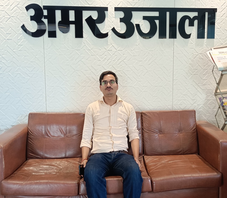 Man seated on a brown leather sofa in an office lobby; large Hindi text on the wall behind him.