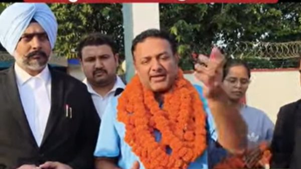 Man in a blue shirt with orange garlands addressing the media, flanked by officials outside a police facility.