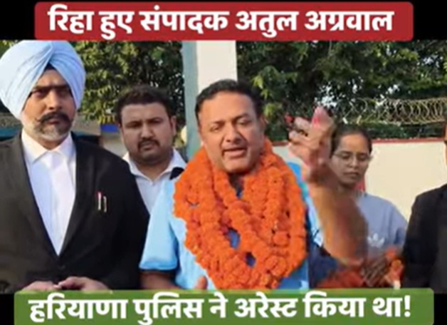 Man in a blue shirt with orange garlands addressing the media, flanked by officials outside a police facility.