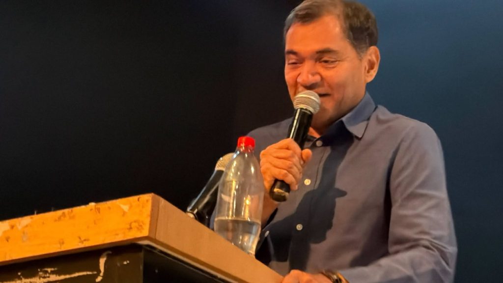 bharat | No. 1 Indian Media News Portal Man in a blue shirt speaking into a microphone at a wooden podium, with a water bottle on the lectern.