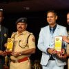 Four men on stage bowing toward the audience as they pose with trophy plaques; two wear tan police uniforms and two wear suits, indicating an award ceremony.