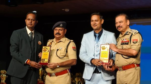 Four men on stage bowing toward the audience as they pose with trophy plaques; two wear tan police uniforms and two wear suits, indicating an award ceremony.