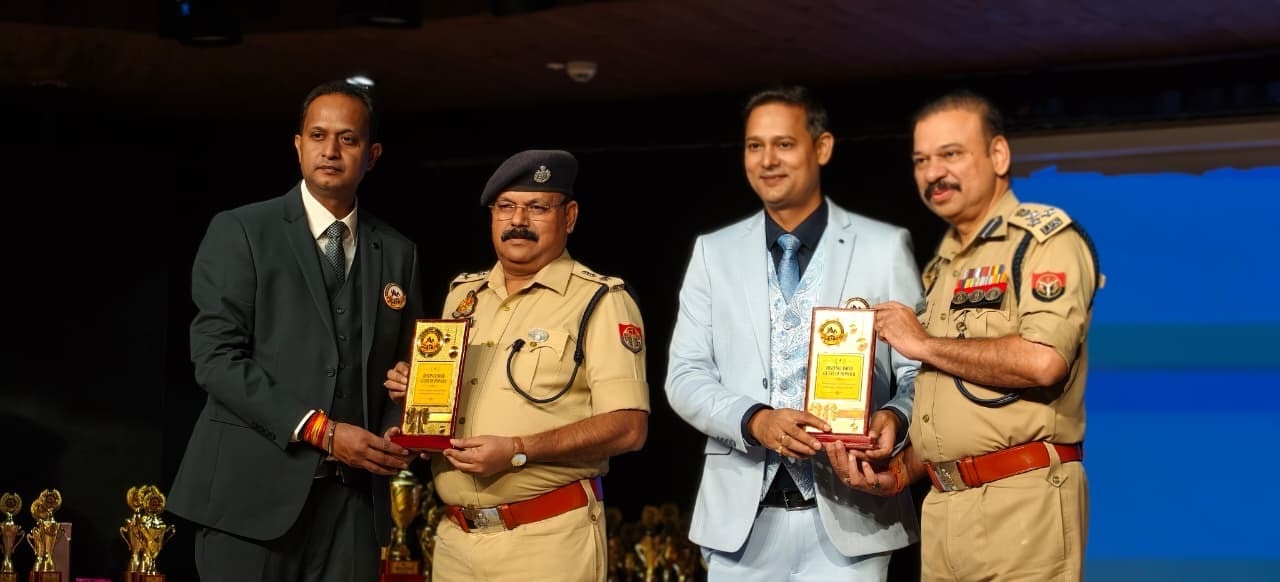 Four men on stage bowing toward the audience as they pose with trophy plaques; two wear tan police uniforms and two wear suits, indicating an award ceremony.