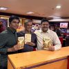 Three men stand at a newsroom counter, each holding a book with a colorful cover and smiling at the camera.