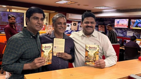 Three men stand at a newsroom counter, each holding a book with a colorful cover and smiling at the camera.