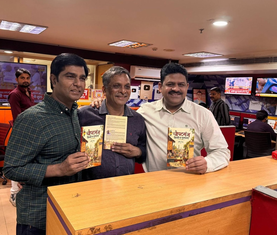 Three men stand at a newsroom counter, each holding a book with a colorful cover and smiling at the camera.