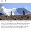 Two hikers walk along a rocky ridge with snowy mountains in the background.
