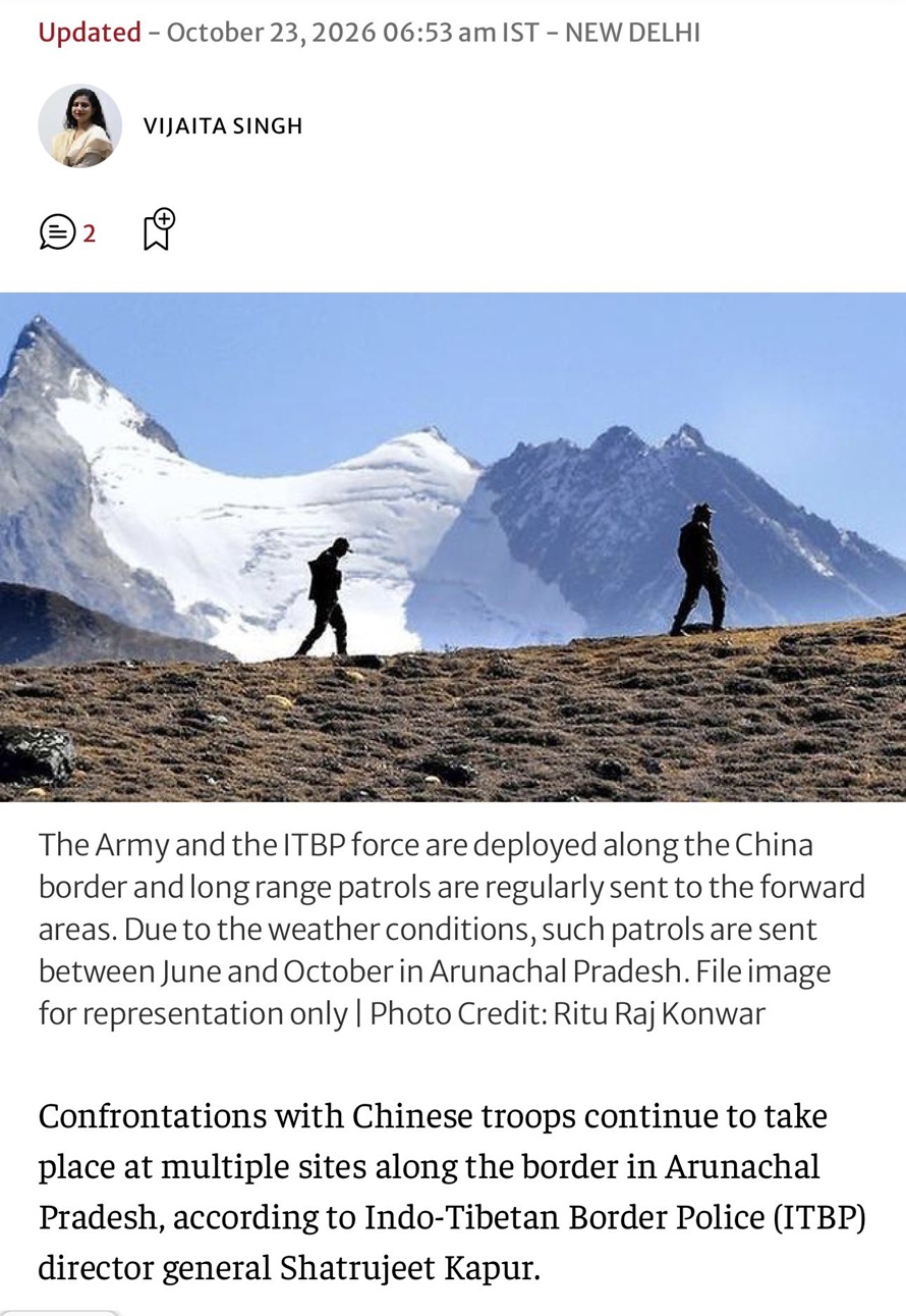 Two hikers walk along a rocky ridge with snowy mountains in the background.