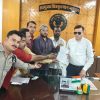 Group of men in an office pose with hands joined over a document, in front of a wooden wall emblem and Hindi sign, Indian flag nearby.