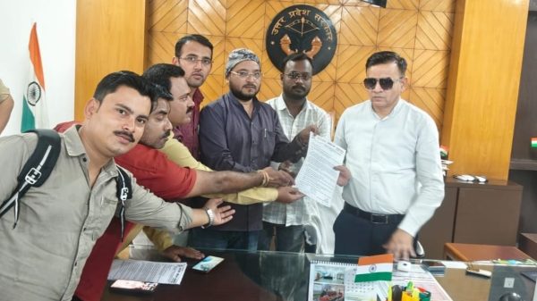 Group of men in an office pose with hands joined over a document, in front of a wooden wall emblem and Hindi sign, Indian flag nearby.