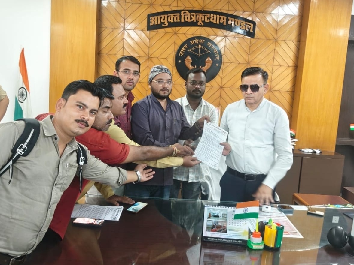 Group of men in an office pose with hands joined over a document, in front of a wooden wall emblem and Hindi sign, Indian flag nearby.