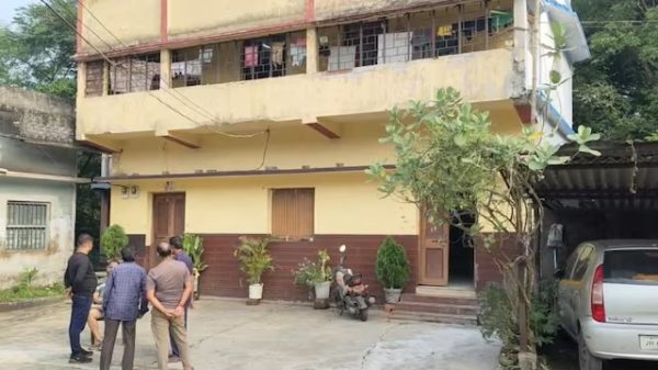Group of men standing and conversing outside a yellow two-story building with potted plants, a scooter, and a parked car on the right.