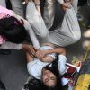 Woman lying on asphalt while several people grab and restrain her arms on a street edge, bystanders nearby.