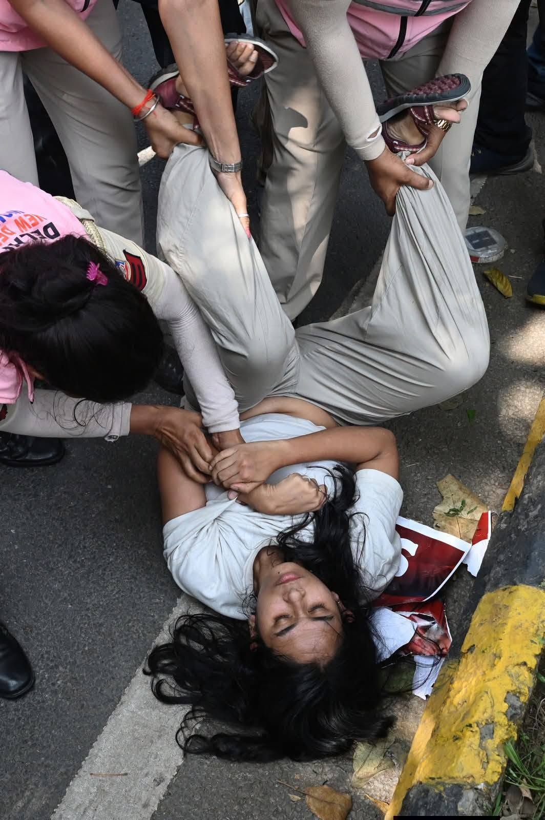 delhi protest | No. 1 Indian Media News Portal Woman lying on asphalt while several people grab and restrain her arms on a street edge, bystanders nearby.