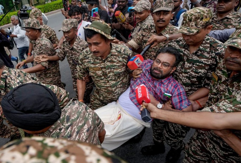 delhi protest_2 | No. 1 Indian Media News Portal Man in a red plaid shirt is restrained by soldiers in camouflage as reporters with microphones crowd around during a tense street scene.