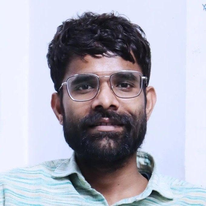 Portrait of a man with dark hair and beard wearing metal-framed glasses and a light green striped shirt, looking at the camera.
