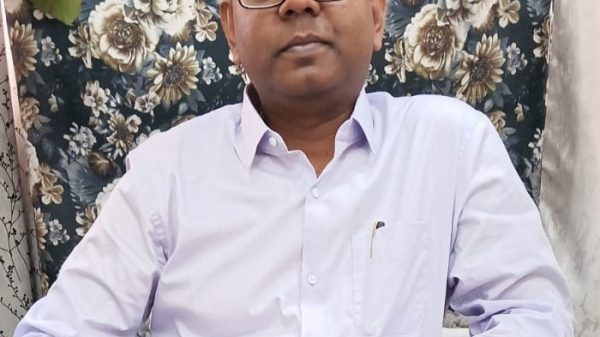 Portrait of a man wearing glasses and a light purple shirt, seated on a floral-patterned chair, with a tilak on his forehead and a wristwatch visible.