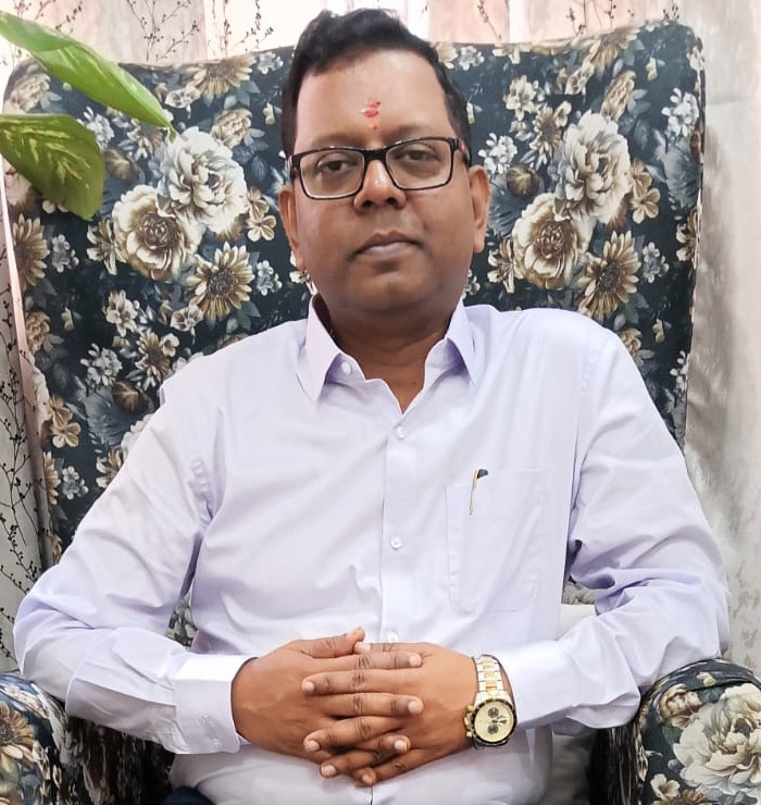 Portrait of a man wearing glasses and a light purple shirt, seated on a floral-patterned chair, with a tilak on his forehead and a wristwatch visible.