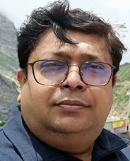 Close-up of a man with dark hair and glasses outdoors, with mountains in the background and a blue jacket.