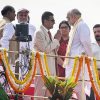 Group of Indian officials on a decorated stage; two men greet with a namaste as garlands drape the railing.
