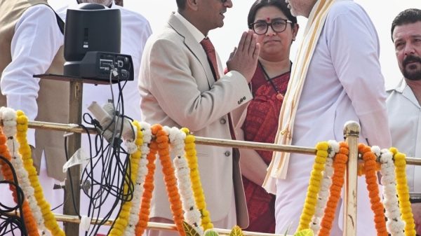 Group of Indian officials on a decorated stage; two men greet with a namaste as garlands drape the railing.