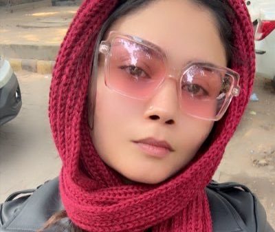 Young woman with a red knit scarf wrapped around her head and neck, wearing pink-tinted oversized sunglasses outdoors.
