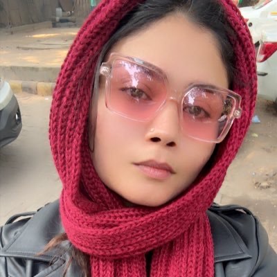 Close-up of a person wearing a red knit scarf and pink-tinted sunglasses outdoors, with parked cars in the background.