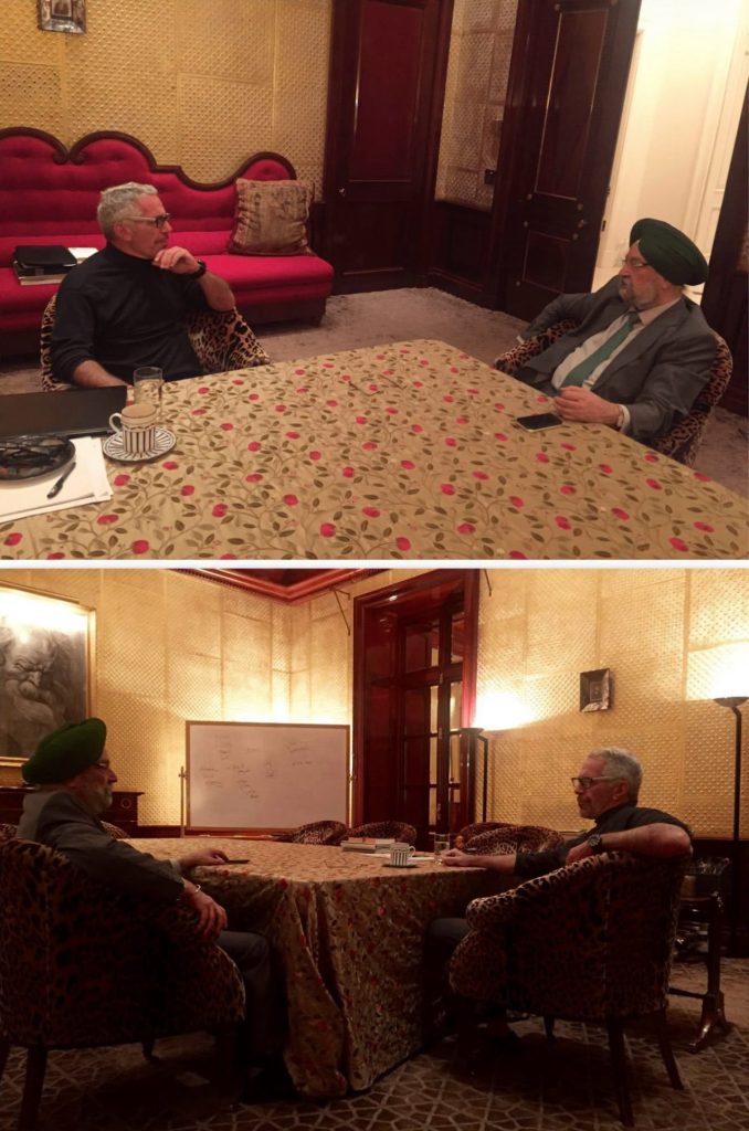 Two older men sit around a round table in a formal lounge, one wearing a green turban and a suit, engaged in conversation.